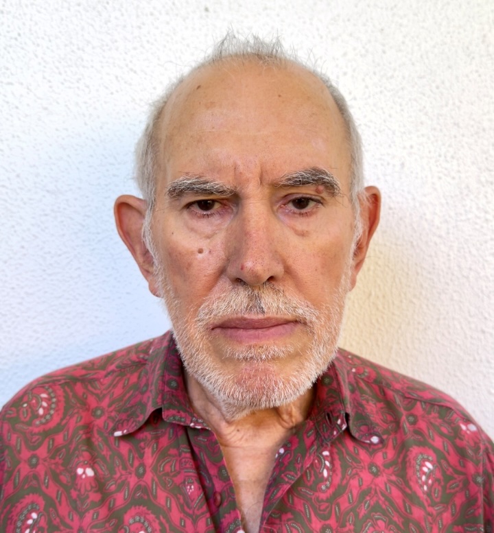 Close-up portrait of an older man with gray hair and beard, wearing a red patterned shirt and looking straight at the camera. Agradecimento: O nosso Serviço Nacional de Saúde