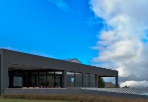 Modern gray building with large glass walls facing the viewer, set against a bright blue sky with clouds.