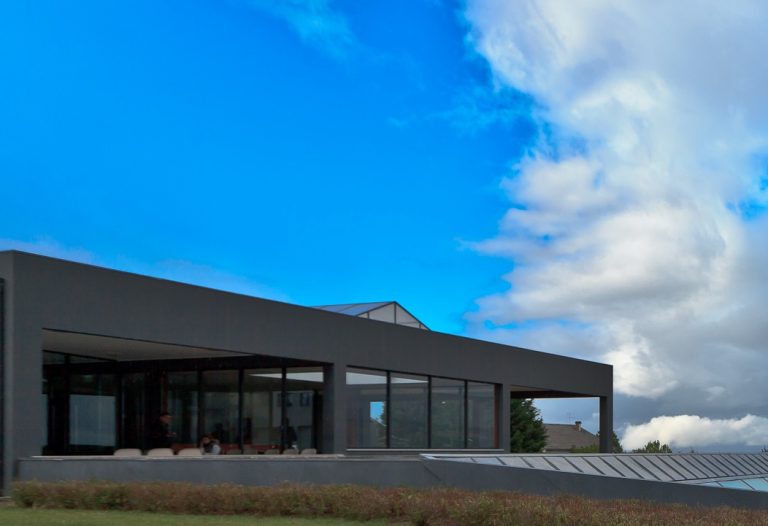 Modern gray building with large glass walls facing the viewer, set against a bright blue sky with clouds.