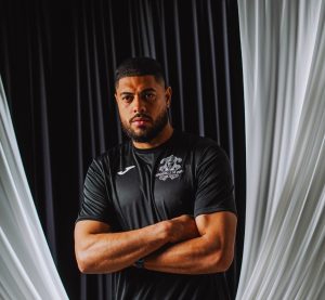 Portrait of a muscular man with arms crossed, wearing a black athletic shirt, standing against dark and light draped curtains.