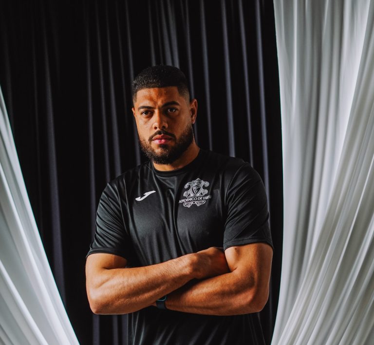 Portrait of a muscular man with arms crossed, wearing a black athletic shirt, standing against dark and light draped curtains.