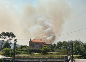 House with orange-tiled roof emitting a thick plume of smoke from a nearby hillside fire, with trees and a yellow railing in the foreground.