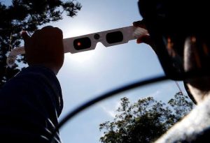 Person holds a white solar eclipse viewing card with two lenses up toward the bright sun, against a blue sky and trees in the background