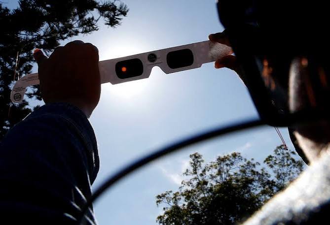 Person holds a white solar eclipse viewing card with two lenses up toward the bright sun, against a blue sky and trees in the background