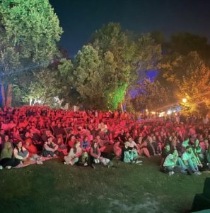 Crowd of people seated on the grass at night under colorful park lights for an outdoor event or concert.
