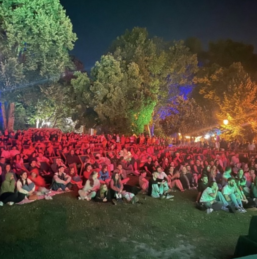 Crowd of people seated on the grass at night under colorful park lights for an outdoor event or concert. Jardim das Artes e das Letras no Parque Aquilino Ribeiro com cinema, concertos, oficinas e futebol