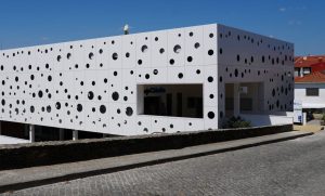 Modern white building with a perforated facade of many black circular holes; cobblestone street in foreground.