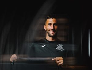 Portrait of a man in a black athletic shirt with a crest, smiling in a dark, moody setting while holding the shirt.