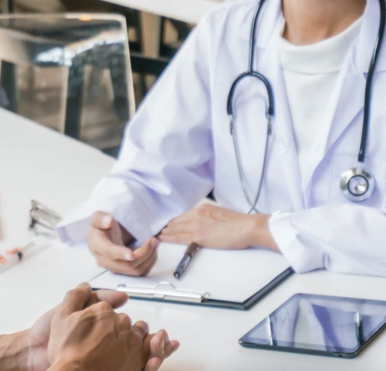 Doctor in a white coat with a stethoscope consults a patient, writing on a clipboard at a desk.