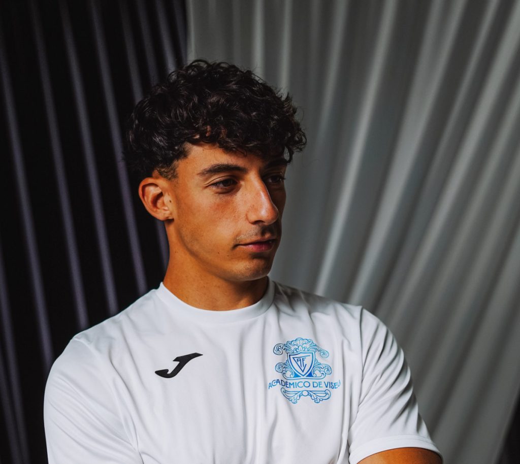 Portrait of a young man in a white sports shirt with a blue crest, looking off to the side against a striped backdrop Académico de Viseu contrata médio espanhol Alejandro Mestanza até 2029
