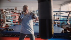 Older man in a white shirt boxing in a gym, fists up and about to punch a hanging bag; boxing ring and equipment visible in the background.