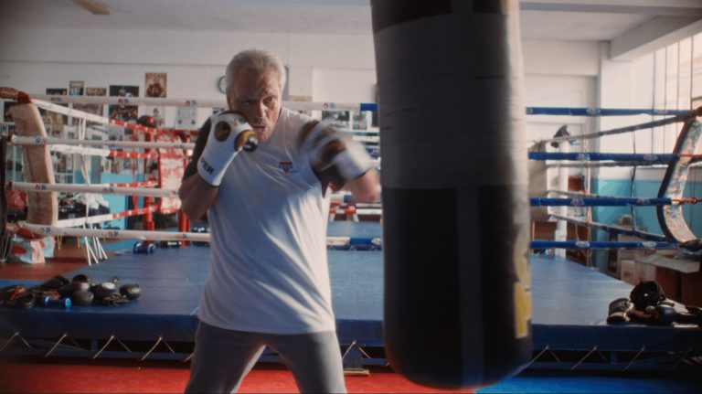 Older man in a white shirt boxing in a gym, fists up and about to punch a hanging bag; boxing ring and equipment visible in the background.