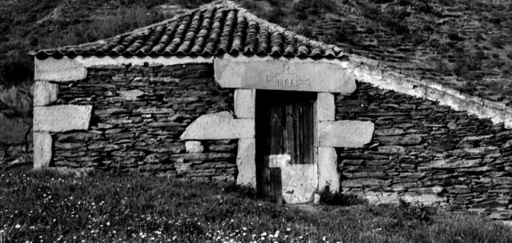 Small stone hut with a tiled roof and a wooden door set in a rough stone wall, in a grassy field. Centro Interpretativo da Mulher Duriense com exposição sobre arquitetura popular do Douro