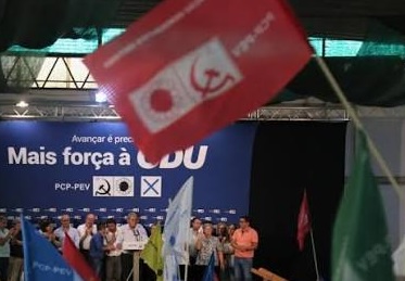 Group of people on a stage at an event; a red flag with a white wheelchair accessibility symbol dominates the foreground, with a blue banner and audience in the background.