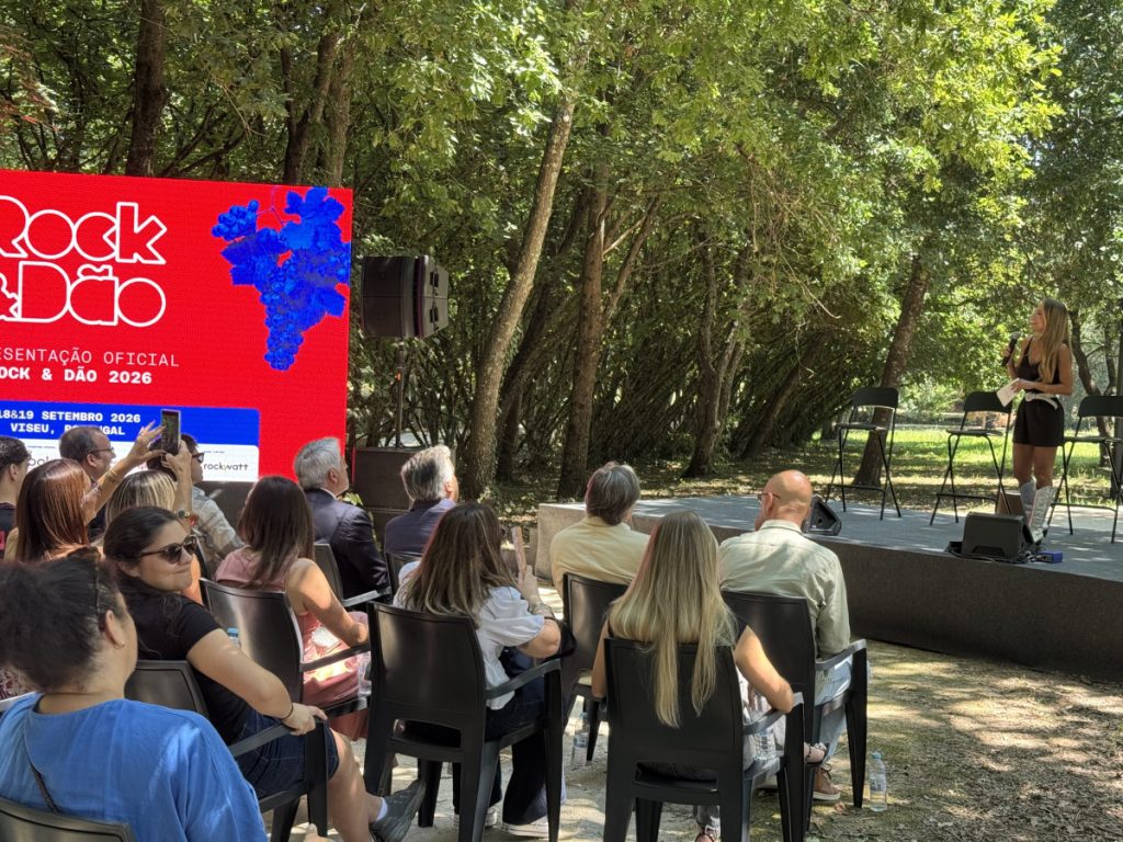 Outdoor stage in a park with a female presenter holding a microphone, addressing a seated audience beneath trees.