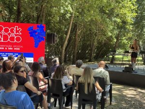 Outdoor stage in a park with a female presenter holding a microphone, addressing a seated audience beneath trees.