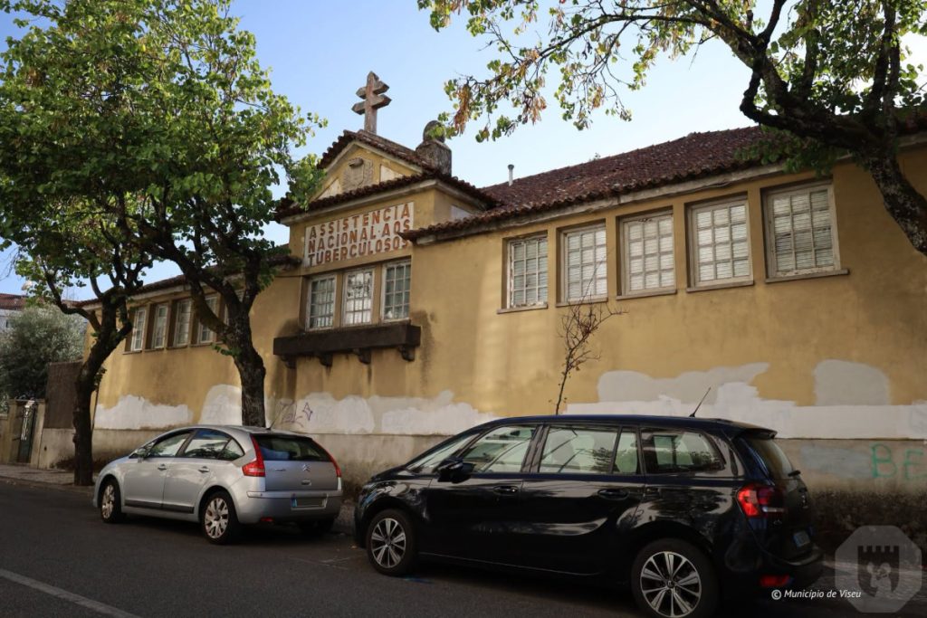 Two cars parked along a street in front of a yellow historic building with a cross on the roof and a sign reading 'Assistência Nacional dos Tuberculosos'. Edifício do Serviço da Luta Anti-Tuberculosa de Viseu acolherá extensão do Museu do Caramulo