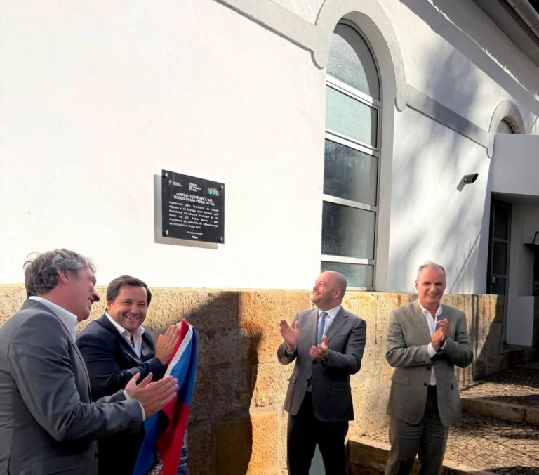 Four men in suits applaud during a plaque unveiling outside a white building with an arched window.