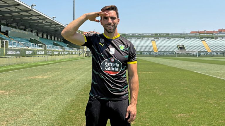 Male soccer player in a black jersey saluting at an empty, sunlit stadium on a green pitch, facing the camera.