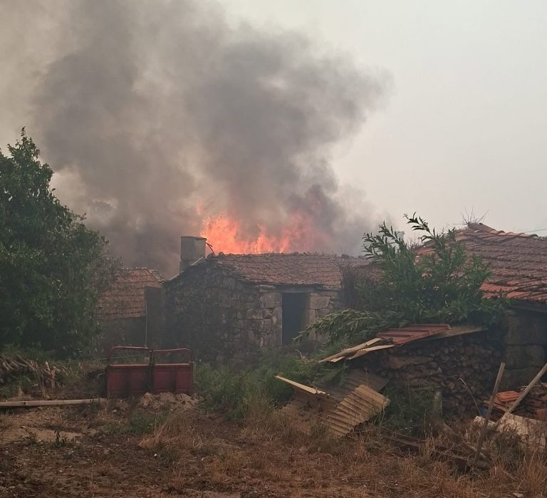 Rural stone houses with red-tiled roofs on fire, large orange flames and thick black smoke billowing upward from the roof.