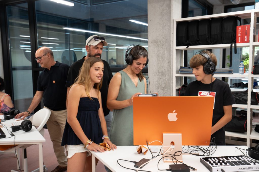 People gathered at a workshop station around an orange iMac; headphones and cables on the desk suggest a tech demo. PPE Tondela assinala primeiro ano do projeto Trilho com mostra de trabalhos de jovens no Centro Tecnológico