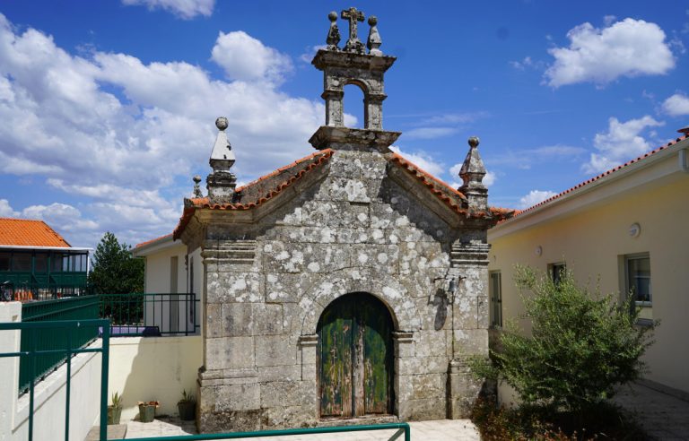 Weathered stone chapel with arched doorway and red-tiled roof, in a sunny courtyard.