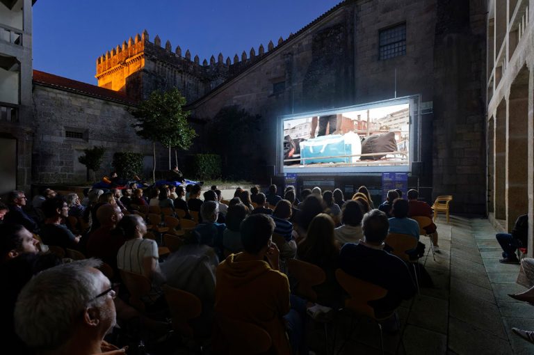 Outdoor cinema in a stone courtyard at night, audience seated watching a large screen.