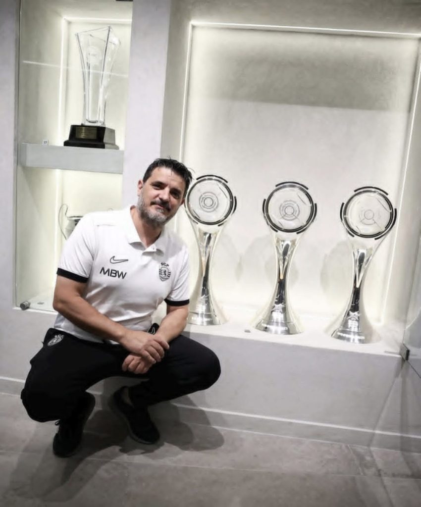 Man in a white polo kneels beside three tall silver trophies displayed in a glass cabinet. Equipa de futsal do Sporting Clube de Portugal estagia em Armamar entre 6 e 9 de agosto
