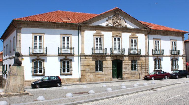 Historic white building with a red tile roof, stone accents, balconies, and a bust statue in the cobblestone plaza.
