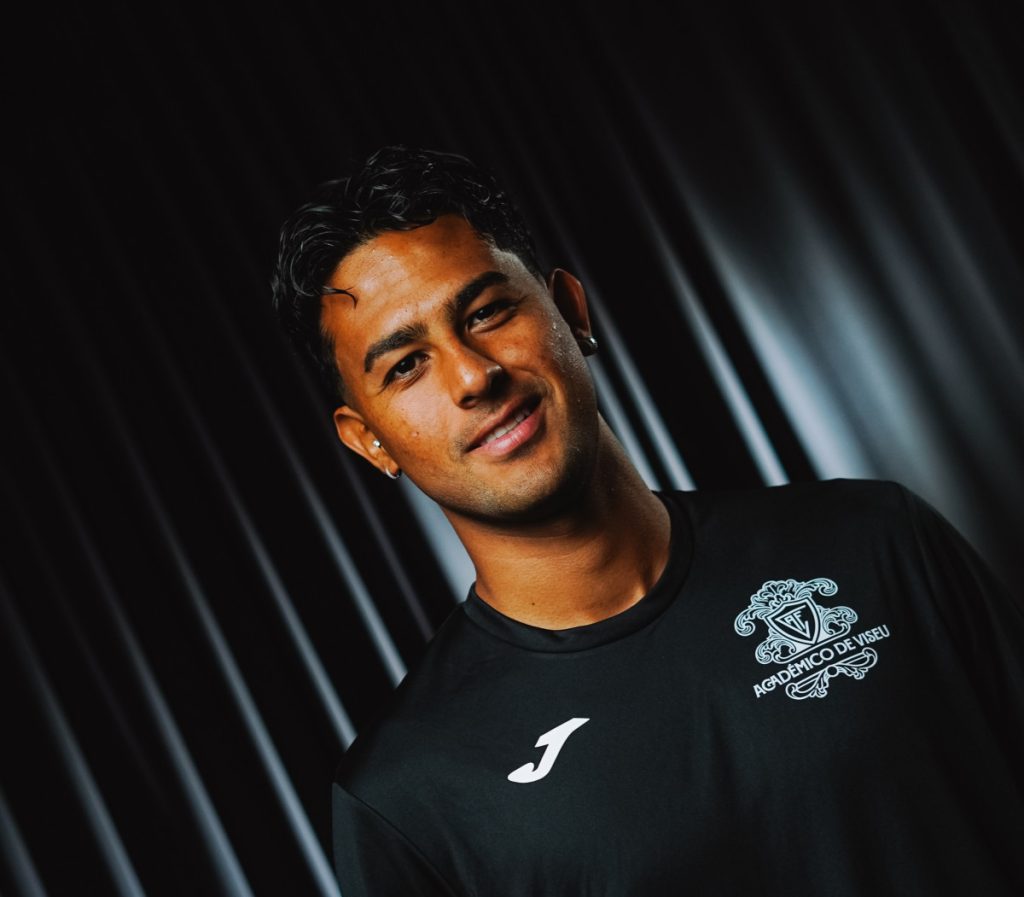 Smiling man in a black athletic shirt with a crest on the chest, angled portrait against a dark striped background. Académico de Viseu garante permanência de Zamora no plantel e primeiro jogo na Luz vai ser sem adeptos