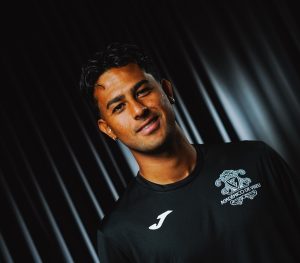 Smiling man in a black athletic shirt with a crest on the chest, angled portrait against a dark striped background.