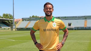Smiling male soccer player in a yellow jersey with green collar on a sunny stadium field, hands on hips.