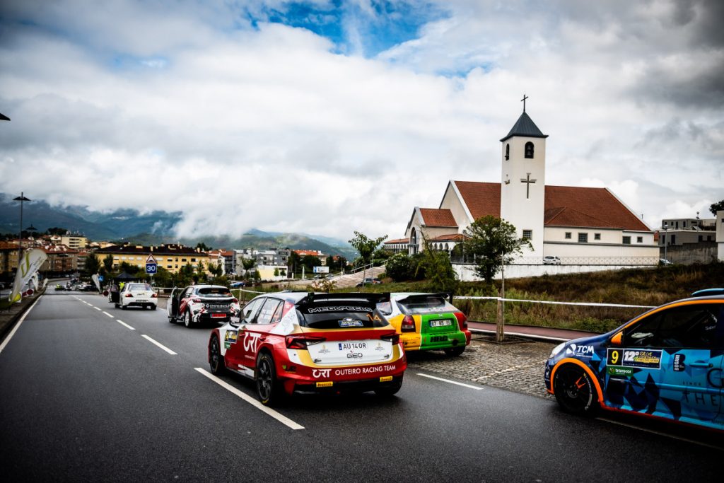 Rally cars parked along a roadway beside a white church with red roof, while a town sits on a hillside under cloudy skies.