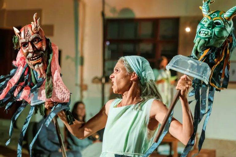 Older woman in a pale green dress performs with two colorful dragon masks on poles indoors.