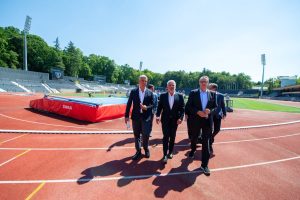 Group of men in suits walking along a red running track beside a padded high-jump mat at an outdoor stadium on a sunny day.