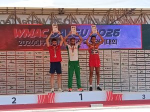Three male athletes stand on a podium holding trophies and wearing medals at an award ceremony, with a WMAC 2026 banner behind them.