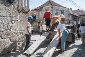 Workers in hard hats guide a small concrete mixer onto a truck using perforated metal ramps on a cobblestone street.