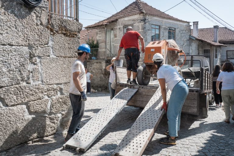 Workers in hard hats guide a small concrete mixer onto a truck using perforated metal ramps on a cobblestone street.