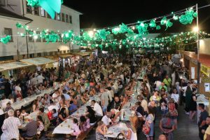 Crowd of people dining at long tables during a nighttime street festival under green decorative lanterns and string lights.