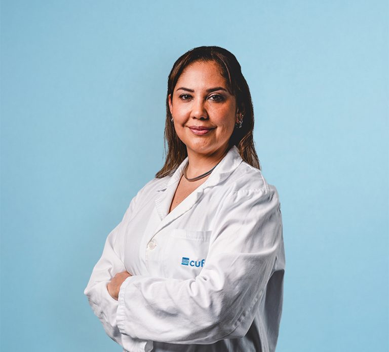 Female clinician in a white coat with arms crossed against a light blue background, looking at the camera.