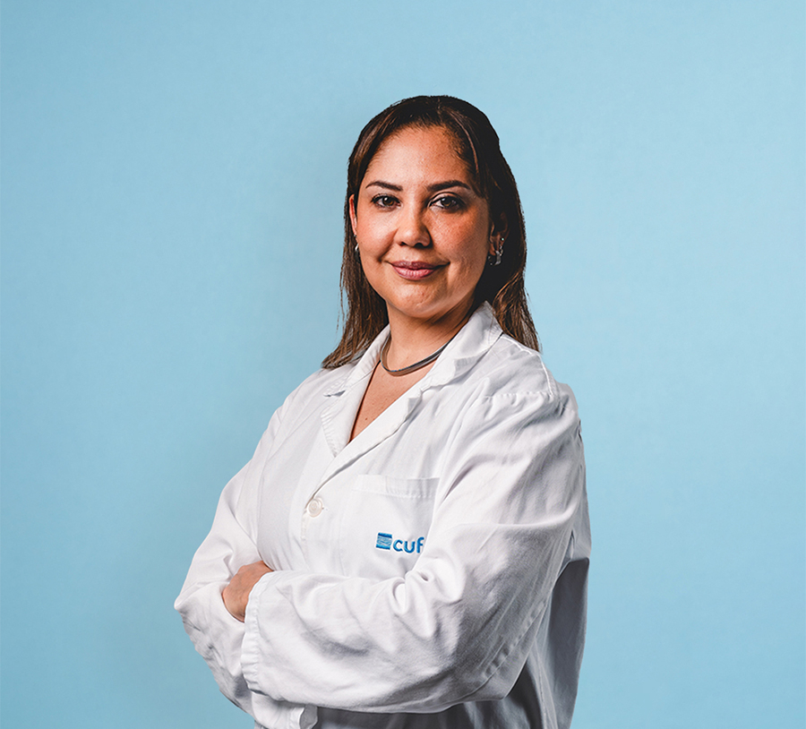 Female clinician in a white coat with arms crossed against a light blue background, looking at the camera.