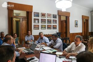 Municipal meeting: diverse group of people sit around a long table with laptops and documents in a wood-paneled room.