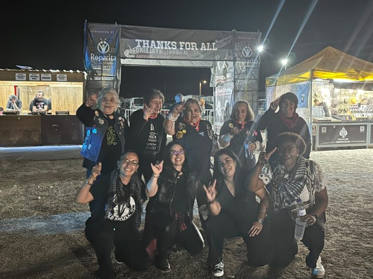 Nine women posing and smiling at a nighttime festival, under a 'THANKS FOR ALL' banner with vendor stalls in the background.