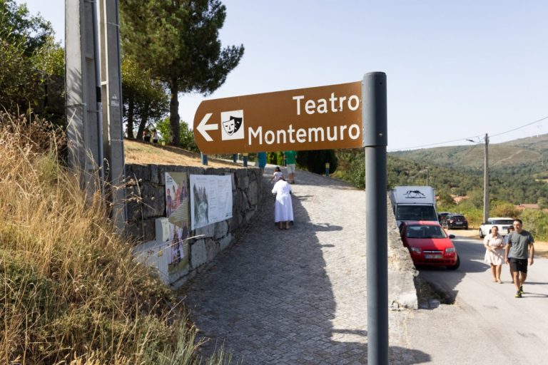 Brown sign reading 'Teatro Montemuro' with a left arrow on a cobbled path; people and parked cars in a rural setting.