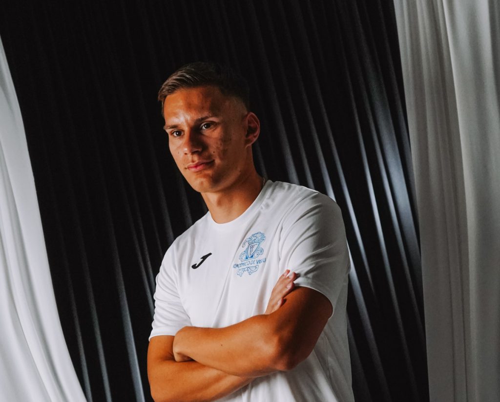 Young man in a white sports jersey with a crest, arms crossed, standing in front of dark curtains. Internacional jovem pela Croácia é reforço do Académico de Viseu
