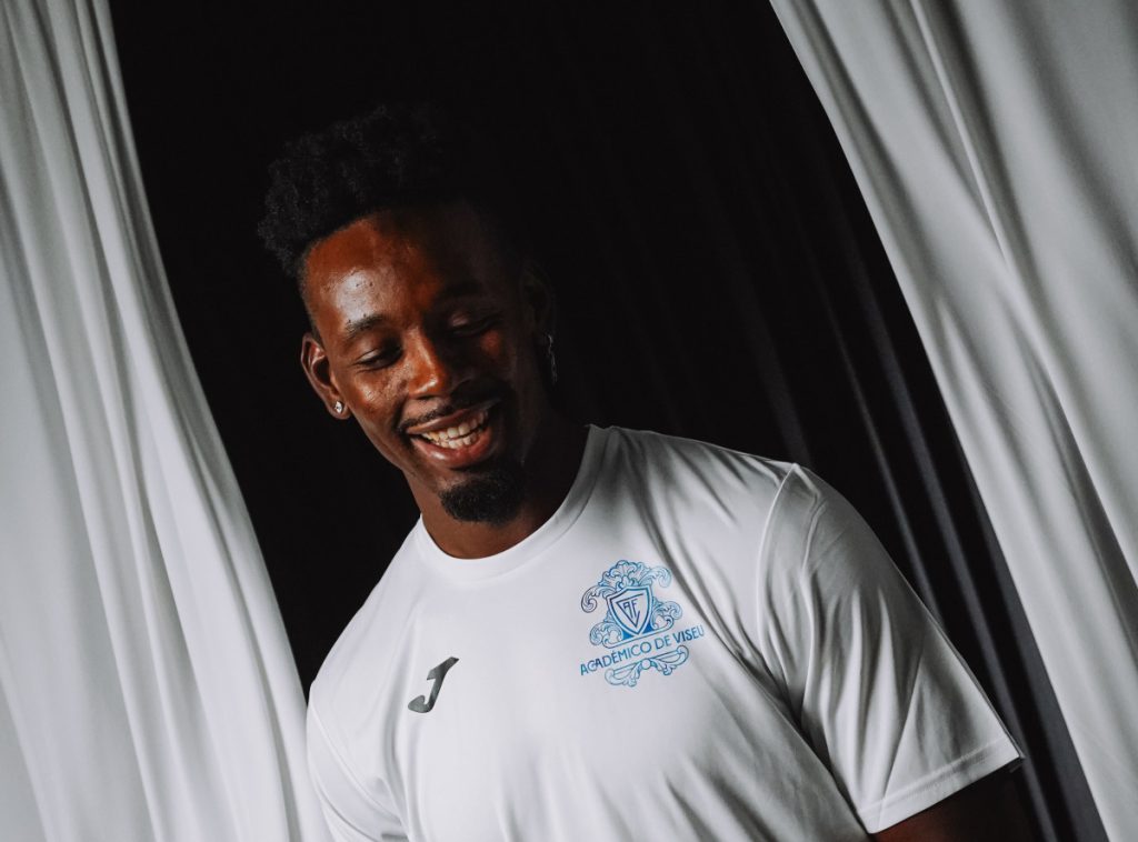 Portrait of a smiling man in a white jersey with a blue Académico de Viseu crest on the chest, standing by white curtains. Avançado marroquino Bouldini regressa a Portugal para jogar no Académico de Viseu