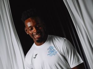 Portrait of a smiling man in a white jersey with a blue Académico de Viseu crest on the chest, standing by white curtains.
