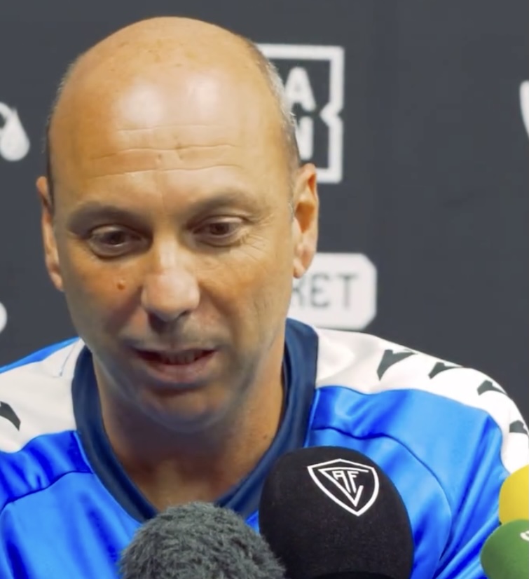 Man in blue sports jersey gives interview, facing microphones in a press setting with a dark backdrop behind him.