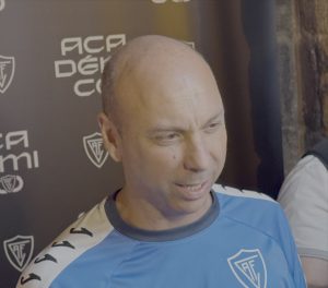 Man in a blue athletic shirt with a team crest being interviewed in front of a branded backdrop.