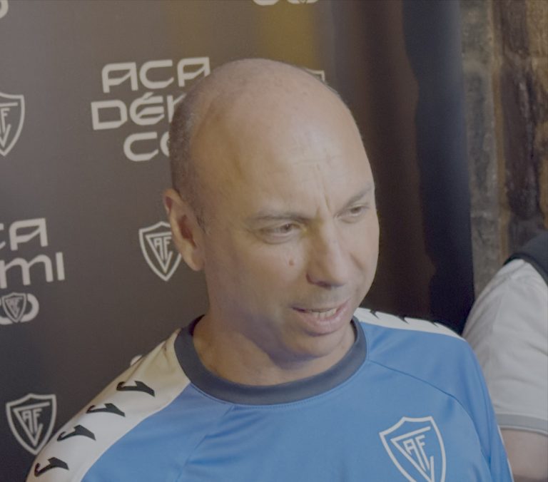 Man in a blue athletic shirt with a team crest being interviewed in front of a branded backdrop.
